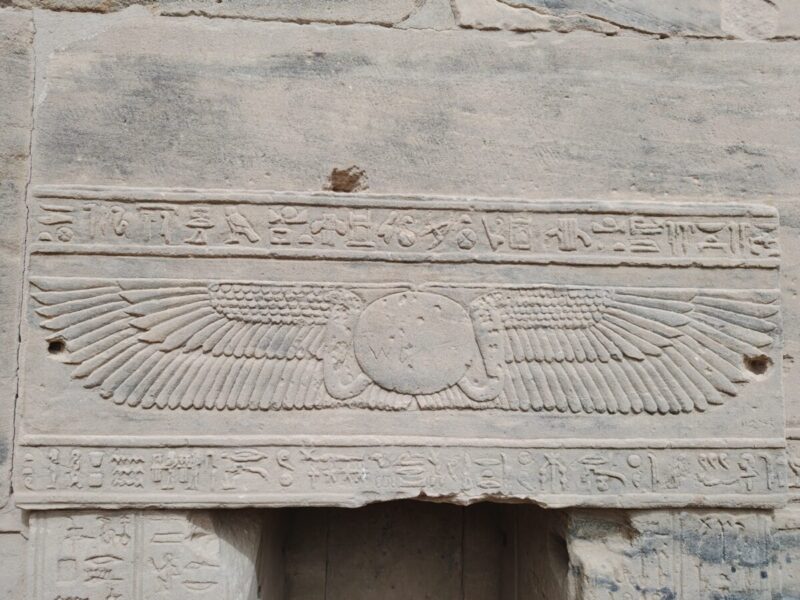 太陽神ラーと天空神ホルスの力が融合した王権の守護印(Winged Sun Disk)。中央の太陽円盤、両側のコブラ、両翼(鷹の翼)がそれぞれ太陽神ラーの力、上下エジプトの統一、ホルス神の守護を表し、神々の力がこの聖域を永遠に守るという意味を持つ象徴です。